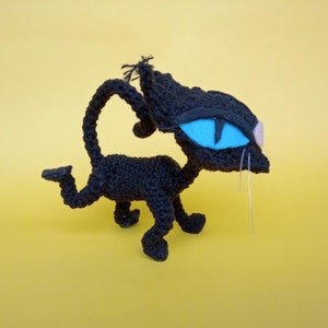 May include: A black crocheted cat toy with blue eyes and white whiskers. The cat is standing on a yellow surface.