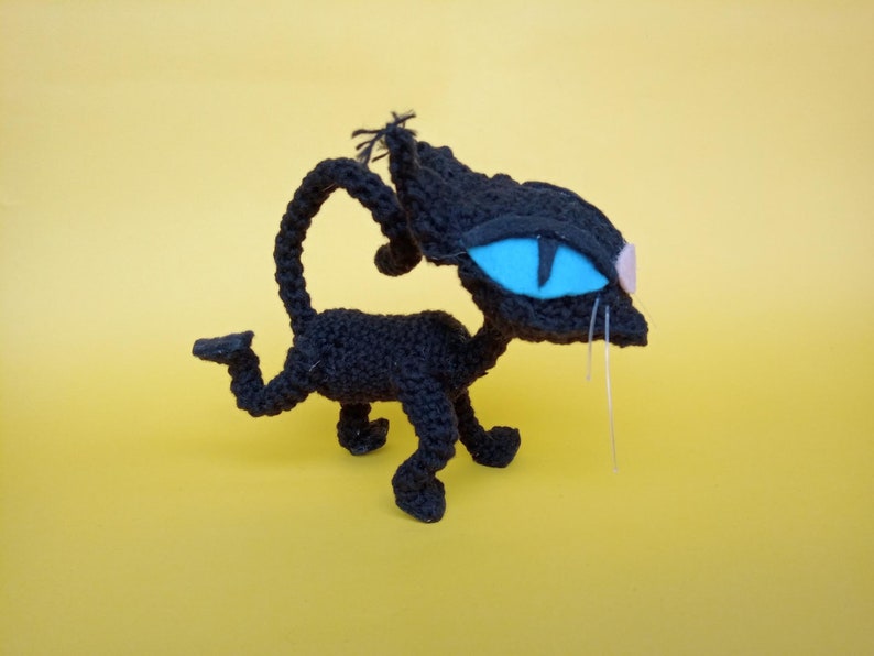 May include: A black crocheted cat toy with blue eyes and white whiskers. The cat is standing on a yellow surface.