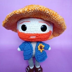 Könnte beinhalten: Eine gehäkelte Puppe, die eine blaue Jacke, einen orangefarbenen Hut und braune Schuhe trägt. Die Puppe hat ein weißes Gesicht mit orangefarbenen Gesichtszügen und schwarzen Augen.