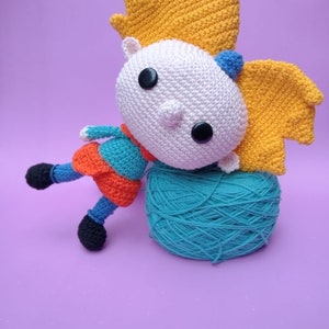 May include: A crocheted doll with yellow hair, blue and orange clothing, and black shoes. The doll is sitting on a ball of teal yarn.