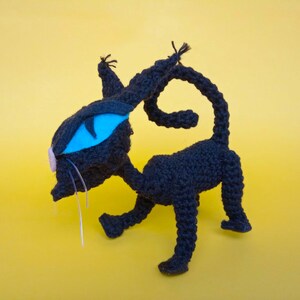May include: A black crocheted cat toy with a blue eye and a pink nose. The cat is standing on a yellow background.