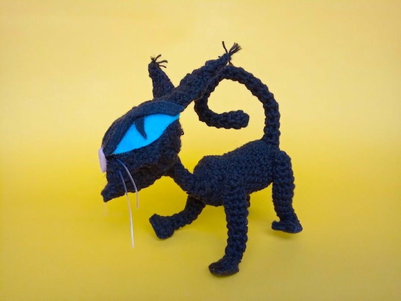 May include: A black crocheted cat toy with a blue eye and a pink nose. The cat is standing on a yellow background.