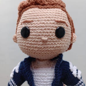 May include: A handmade crochet doll with a light skin tone, brown hair, and black button eyes. The doll wears a navy blue jacket with white accents over a white shirt. The doll's hair is styled in a textured, curly fashion.