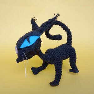 May include: A black crocheted cat toy with a blue eye and a pink nose. The cat is standing on a yellow surface.