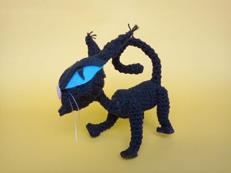 May include: A black crocheted cat toy with a blue eye and a pink nose. The cat is standing on a yellow surface.