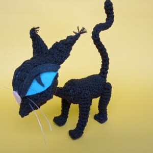 May include: A black crocheted cat toy with blue eyes and a pink nose. The cat is standing on a yellow surface.