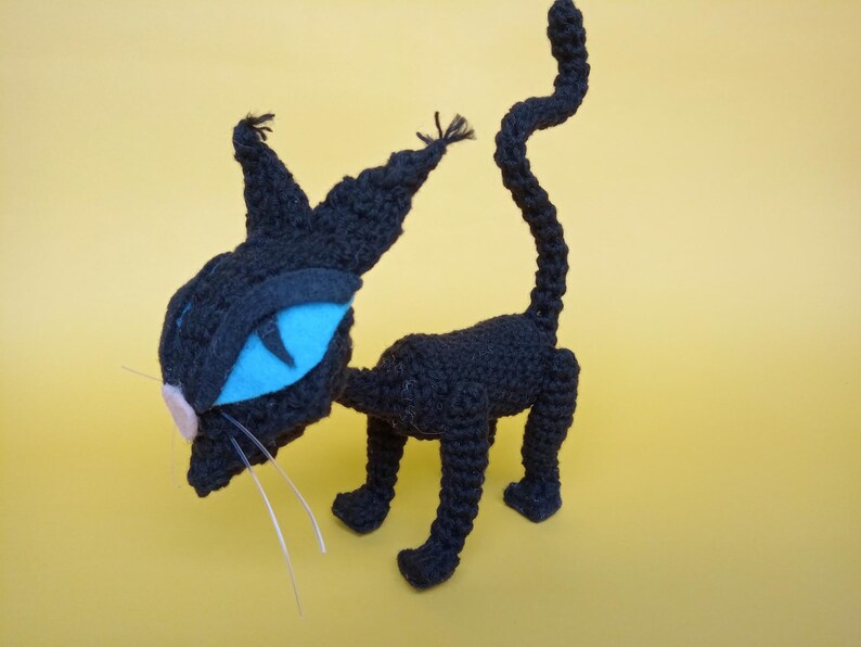 May include: A black crocheted cat toy with blue eyes and a pink nose. The cat is standing on a yellow surface.