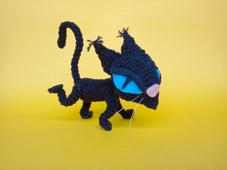 May include: A black crocheted cat toy with blue eyes and a pink nose. The cat is in a walking pose with its tail curled up.