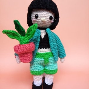 Puede incluir: Muñeca de ganchillo de un personaje con un corte bob negro, chaqueta verde azulado, top negro y pantalones cortos verdes. La muñeca sostiene una maceta rosa con una planta verde. La muñeca tiene zapatos negros y calcetines blancos.