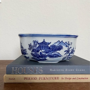 May include: A white ceramic bowl with a blue transferware pattern featuring pagodas, trees, and mountains. The bowl has a scalloped rim and sits on a stack of two books, one titled HOUSES by Mariette Himes Gomez and one titled PERIOD FURNITURE.