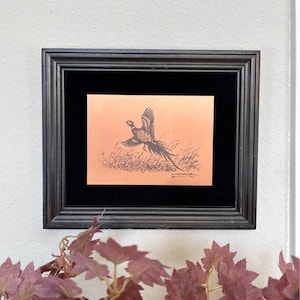 May include: A rectangular black frame containing a copper-colored print of a pheasant in flight over tall grass. The artwork features a black ink illustration of the bird and includes a signature in the bottom right corner, set against a black mat.