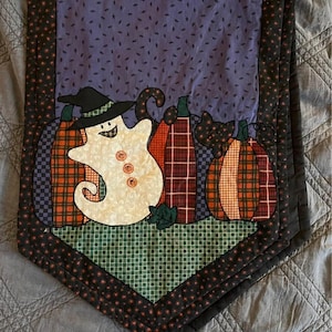 May include: A quilted table runner featuring a central purple panel with a ghost wearing a black hat, three plaid pumpkins in orange, red, and green, and a black cat silhouette. The border is black with small red stars and a green checkered base.