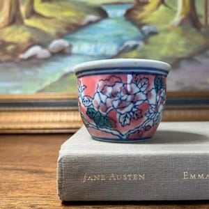 May include: A small, hand-painted ceramic cup with a floral design. The cup has a white rim, a pink body with white and blue floral patterns, and a blue base. The cup is on a book with the title "Jane Austen" visible.