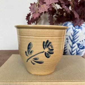 Puede incluir: Una vasija de cerámica color canela con un diseño floral pintado a mano en azul oscuro en la parte frontal. El recipiente tiene una forma cónica con un borde ancho y una hendidura horizontal cerca de la parte superior. Superficie lisa.