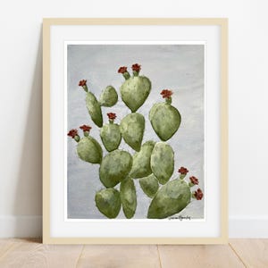 Puede incluir: Pintura a la acuarela de un cactus verde con flores rojas, sobre un fondo gris claro. La obra está enmarcada en un marco de madera clara, adecuado para la decoración del hogar. La firma del artista es visible en la esquina inferior derecha.