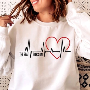 May include: White sweatshirt featuring a black and red heart rate graphic and the text "THE BEAT GOES ON". The design is a casual style.