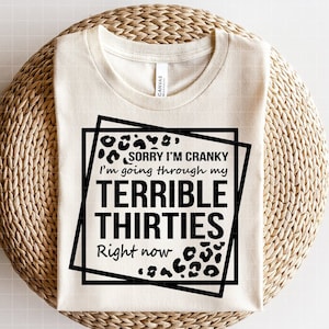 Könnte beinhalten: Cremefarbenes T-Shirt mit dem schwarzen Text "SORRY I'M CRANKY I'm going through my TERRIBLE THIRTIES Right now" mit Leopardenmuster-Akzenten. Das Shirt wird auf einer gewebten, kreisförmigen Oberfläche präsentiert.