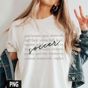 May include: White t-shirt with the word "soccer" written in cursive and a list of soccer terms below it, including "goal keeper", "pass", "footwork", "half field", "cross ball", "kick", "header", "defender", "sweeper", "penalty", "goal line", "breakaway", "control", "teamwork", and "angles".