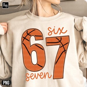 May include: A beige sweatshirt featuring the numbers "67" in a basketball design, with the word "six" above and "seven" below in a handwritten font. The basketball design is orange with black lines. The sweatshirt is a neutral color.
