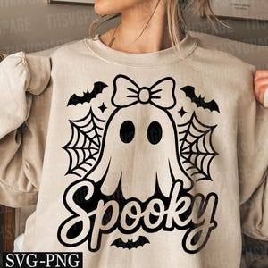 May include: Beige sweatshirt with a black graphic of a ghost wearing a bow, spiderwebs, bats, and the word "Spooky". The design is perfect for Halloween and fall. The sweatshirt is made of a soft material.