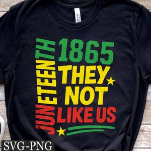 May include: Black t-shirt featuring the words "JUNETEENTH 1865 THEY NOT LIKE US" in green, yellow, and red. The design includes stars and green lines. A white and black trainer is visible in the lower left corner.