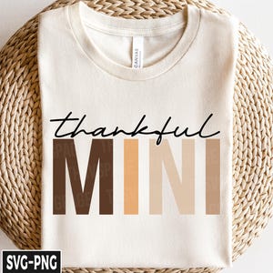 May include: Cream-colored t-shirt with the words "thankful MINI" in black script and block letters. The word "MINI" is in shades of brown and beige. The shirt is displayed on a woven basket.