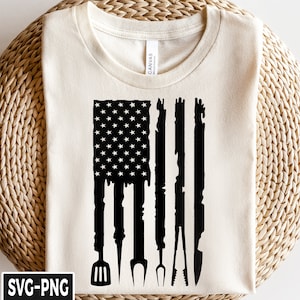 Puede incluir: Camiseta color crema con un diseño gráfico negro de la bandera estadounidense. Las rayas de la bandera están estilizadas como utensilios de barbacoa, incluyendo una espátula, un tenedor y unas pinzas. La camiseta tiene cuello redondo y mangas cortas.