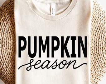 May include: Cream-colored t-shirt with the words "PUMPKIN season" printed in black. The word "PUMPKIN" is in a bold, sans-serif font, while "season" is in a cursive script. The shirt is displayed on a woven surface.