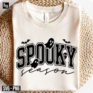 Puede incluir: Camiseta color crema con las palabras "SPOOKY season" en negro, con gr&aacute;ficos de fantasmas y murci&eacute;lagos. El texto tiene una fuente de estilo universitario. La camiseta est&aacute; doblada sobre una superficie tejida de color marr&oacute;n claro.