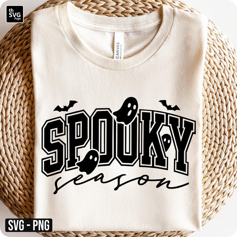 Puede incluir: Camiseta color crema con las palabras "SPOOKY season" en negro, con gr&aacute;ficos de fantasmas y murci&eacute;lagos. El texto tiene una fuente de estilo universitario. La camiseta est&aacute; doblada sobre una superficie tejida de color marr&oacute;n claro.