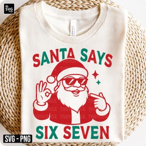 Könnte beinhalten: Cremefarbenes T-Shirt mit festlichem Design. Die Grafik zeigt den Weihnachtsmann mit Sonnenbrille und den Text "SANTA SAYS SIX SEVEN" in Rot und Grün. Das Shirt besteht aus weichem Material und ist perfekt für die Weihnachtszeit.