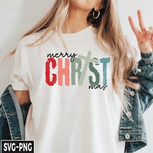 May include: A white t-shirt with the text "merry CHRISTmas" in colorful, brushstroke lettering. A green and blue cross is centered between the words "CHRIST" and "mas".