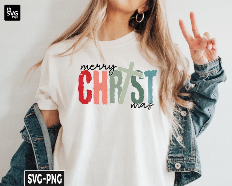 May include: A white t-shirt with the text "merry CHRISTmas" in colorful, brushstroke lettering. A green and blue cross is centered between the words "CHRIST" and "mas".