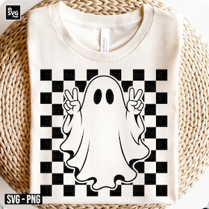 Op de afbeelding: Een crèmekleurig t-shirt met een zwart-wit geblokt patroon op de voorzijde. Gecentreerd over het patroon staat een witte geestillustratie met zwarte omlijning, afgebeeld met beide handen omhoog in een vredesteken.