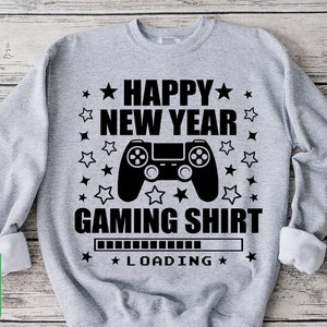 K&ouml;nnte beinhalten: Ein graues Sweatshirt mit einem schwarzen Grafikdesign eines Videospielcontrollers und dem Text "Happy New Year Gaming Shirt" mit einem Ladebalken darunter. Der Text ist von schwarzen Sternen umgeben.