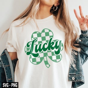 May include: A white t-shirt with a green shamrock design. The shamrock is made up of checkered squares in green and white. The word "Lucky" is written in green cursive letters inside the shamrock.