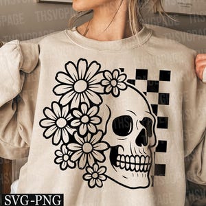 May include: Beige sweatshirt featuring a black and white floral skull design. The skull is on the right side, with a checkerboard pattern behind it. The design includes daisies and the text "SVG-PNG".