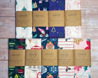 May include: A collection of reusable paperless towels with various Christmas-themed patterns. Designs include Christmas trees, ornaments, bows, and floral prints in red, green, blue, and white. Each towel is wrapped with a brown paper band.