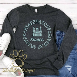 May include: A dark gray long-sleeve shirt with a teal blue circle graphic. The graphic features a building with a dome and the text "Beauxbatons Academy of Magic* France".