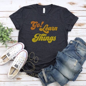 Puede incluir: Una camiseta gris oscuro con el texto "Go! Learn Things" en una fuente retro naranja y amarilla.