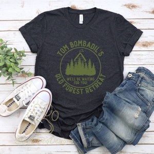 May include: A dark gray t-shirt with a green circle graphic that says "Tom Bombadil's Old Forest Retreat" with a cabin and trees inside the circle. The text "We'll be waiting for you!" is inside the circle.