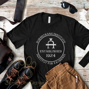 May include: Black t-shirt with a white graphic of the Durmstrang Institute logo. The logo features a stylized letter "A" with a diamond in the center, surrounded by a circle with the text "Durmstrang Institute Established 1924 Institut für Zauberei".