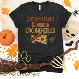 Op de afbeelding: Een donkergrijs T-shirt met de tekst "Autumn Leaves & Murder Documentaries" in oranje en bruine letters. De tekst wordt omringd door een bloemmotief met rode, oranje, gele en groene bloemen.