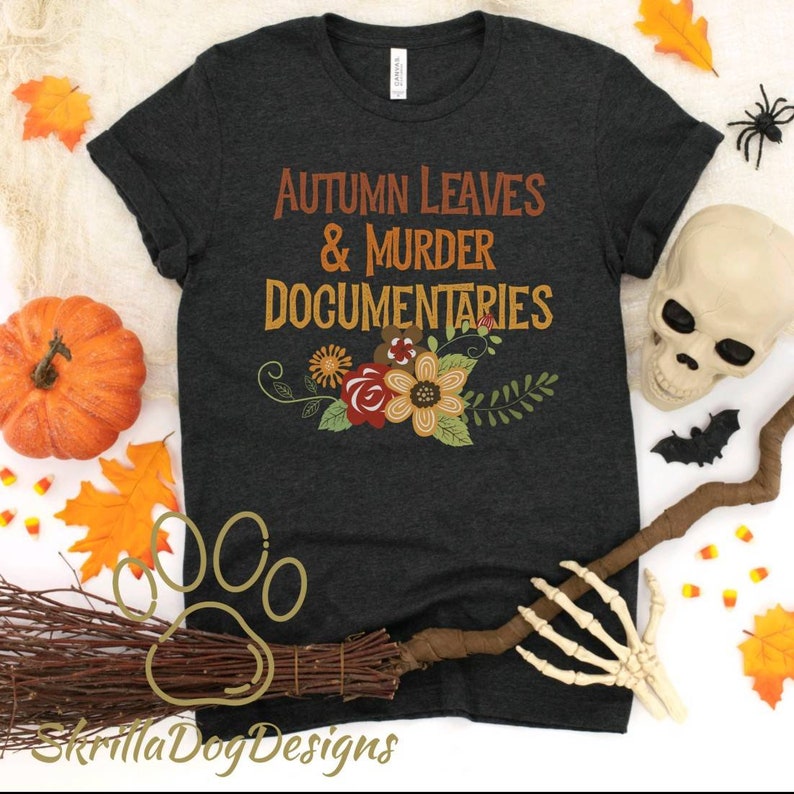 Op de afbeelding: Een donkergrijs T-shirt met de tekst "Autumn Leaves & Murder Documentaries" in oranje en bruine letters. De tekst wordt omringd door een bloemmotief met rode, oranje, gele en groene bloemen.