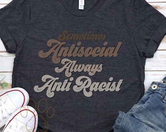 Puede incluir: Una camiseta gris oscuro con el texto "Sometimes Antisocial Always Anti Racist" en letras marrones y beige con un aspecto vintage desgastado.