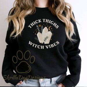 May include: Black sweatshirt with the text "Thick Thighs Witch Vibes" in white lettering. The design features a cluster of crystals with a crescent moon and stars. A paw print is visible in the bottom left corner.