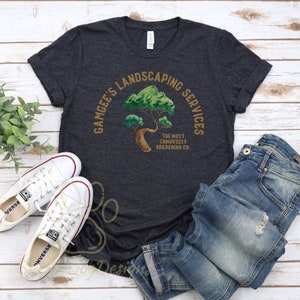 May include: A dark gray t-shirt with a gold circle design that says "Gamgee's Landscaping Services" and features a green tree with the text "The Most Famousest Gardening Co."