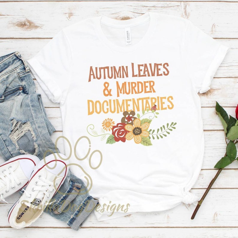 Op de afbeelding: Wit T-shirt met de tekst "Autumn Leaves & Murder Documentaries" in oranje en bruine letters. De tekst wordt omringd door een bloemmotief met rode, oranje, gele en groene bloemen.