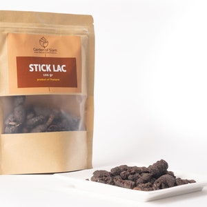 Può includere: Un sacchetto di carta marrone con una finestra di plastica trasparente che contiene lacca in bastoncini marrone scuro, una resina naturale. Il sacchetto ha un'etichetta che recita "Garden of Siam" e "Stick Lac 100 gr prodotto della Thailandia". In primo piano c'è un piccolo piatto bianco con un mucchio di lacca in bastoncini.