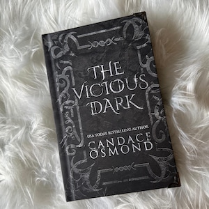 May include: A black hardcover book with a silver design on the cover. The title of the book is "The Vicious Dark" by Candace Osmond.